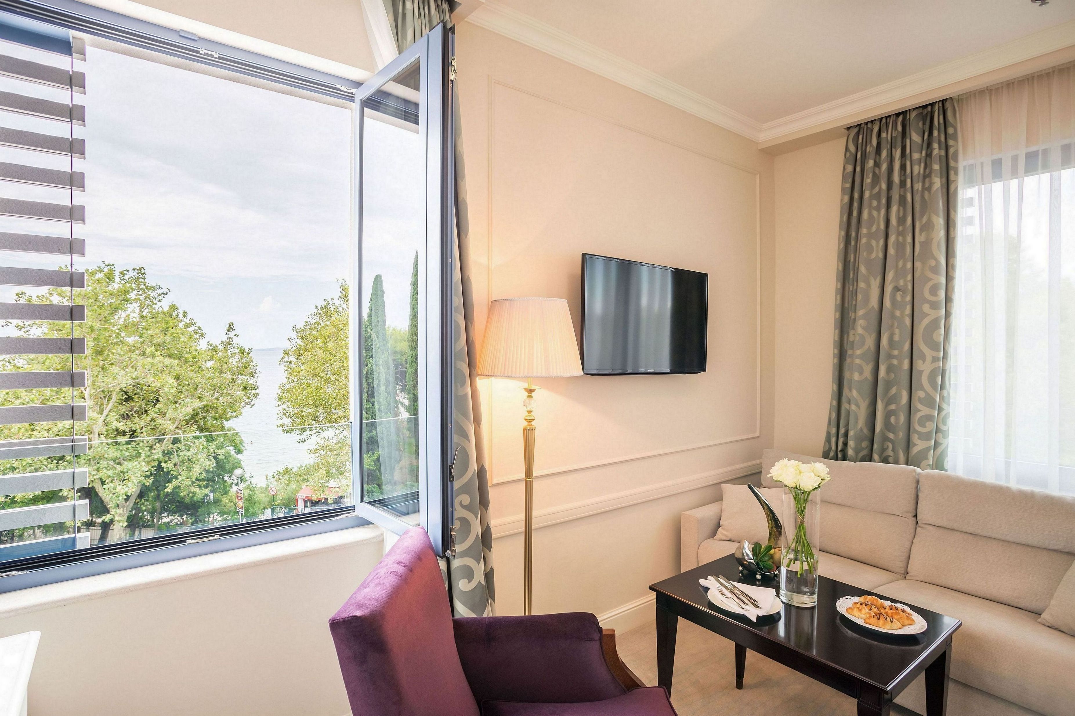 A hotel room with a large window, a sofa, a TV mounted on the wall, and a small table with a vase of flowers.