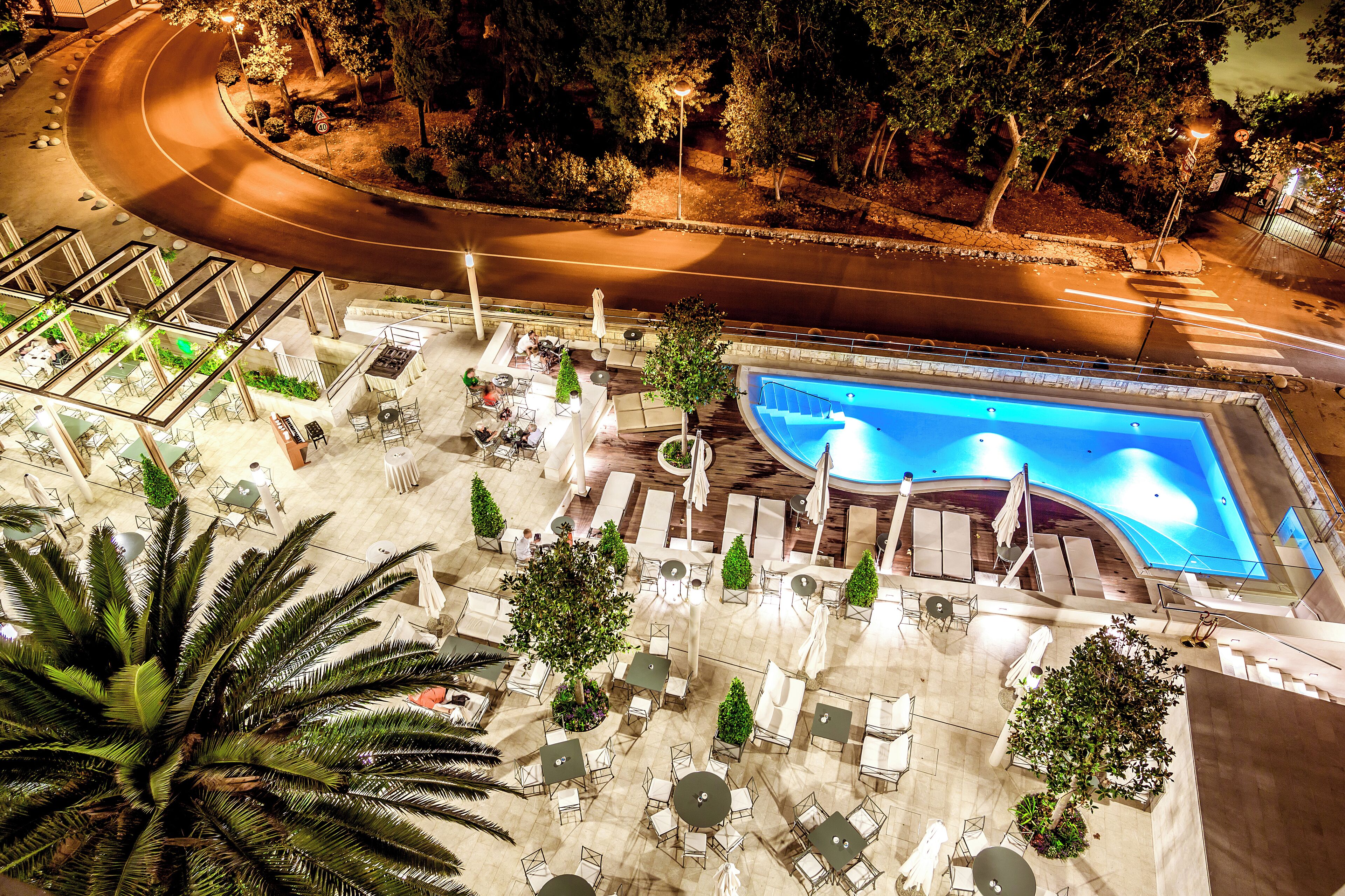 An outdoor pool area with lounge chairs and tables, surrounded by trees and a curved pathway.