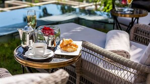 A table set with a coffee cup, a glass of wine, a teapot, a glass of water, and croissants on a wicker chair overlooking a pool. - Hotel Park (Split)