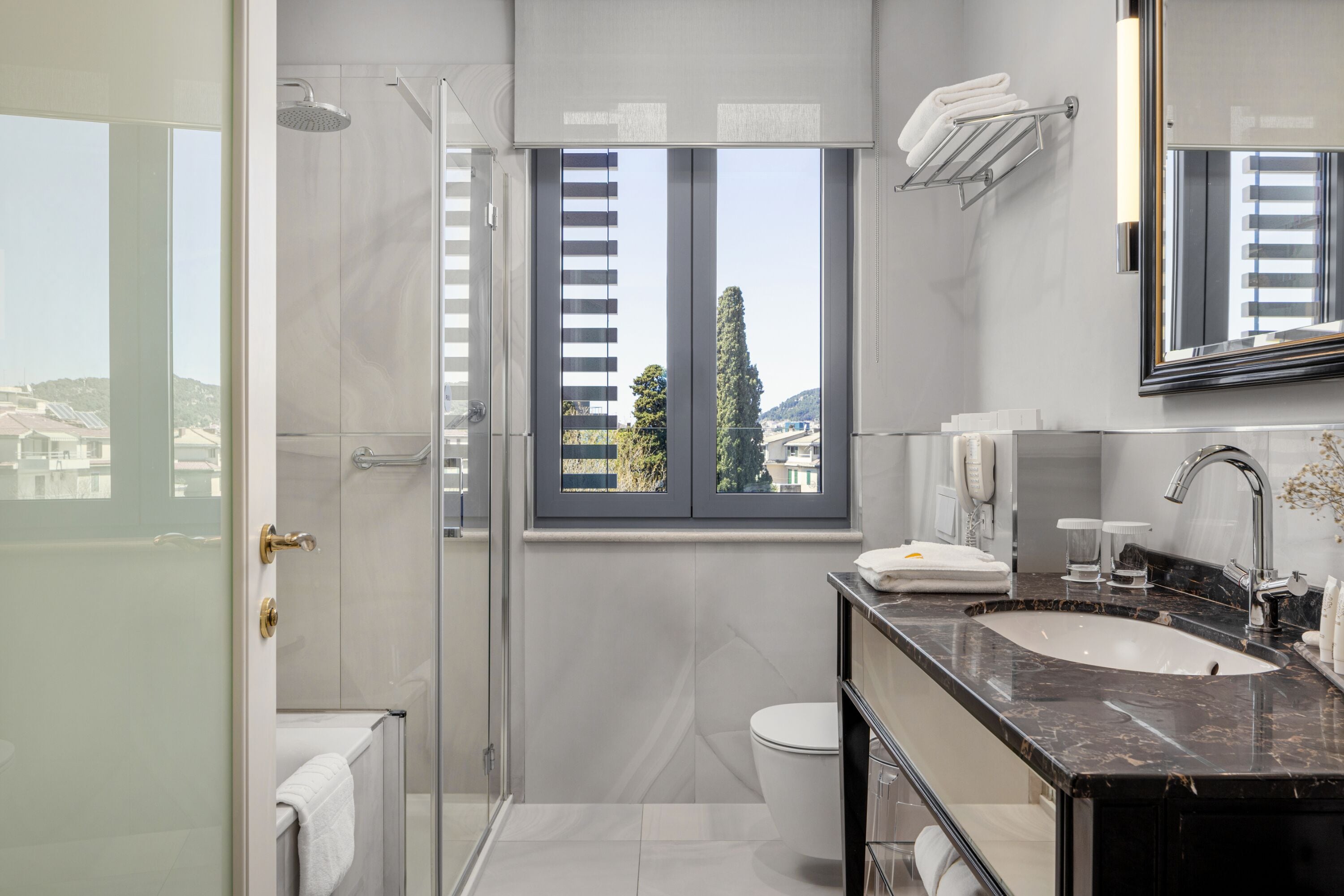 A modern bathroom with a glass shower enclosure, a large window with blinds, a sink with a marble countertop, and a toilet.