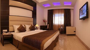 Quarto de hotel com uma cama grande, uma escrivaninha, uma cadeira, uma televisão e um abajur.