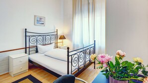 Une chambre à coucher bien rangée, avec un lit en métal, une table de chevet avec une lampe, une petite table d’appoint, une chaise et une plante en pot ornée de fleurs.