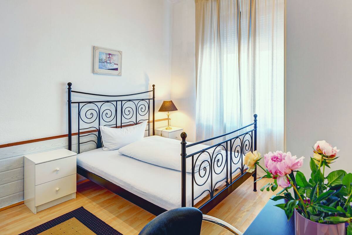 A neatly arranged bedroom with a metal bed frame, a bedside table with a lamp, a small bedside table, a chair, and a potted plant with flowers.