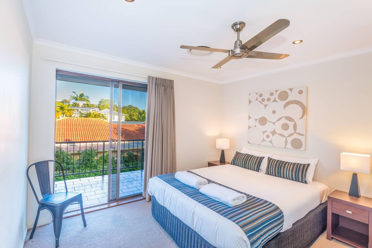A hotel room with a bed, a chair, a ceiling fan, a nightstand, and a view of the outside.