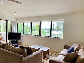 A living room with a TV, two sofas, a coffee table, and a view of greenery outside.