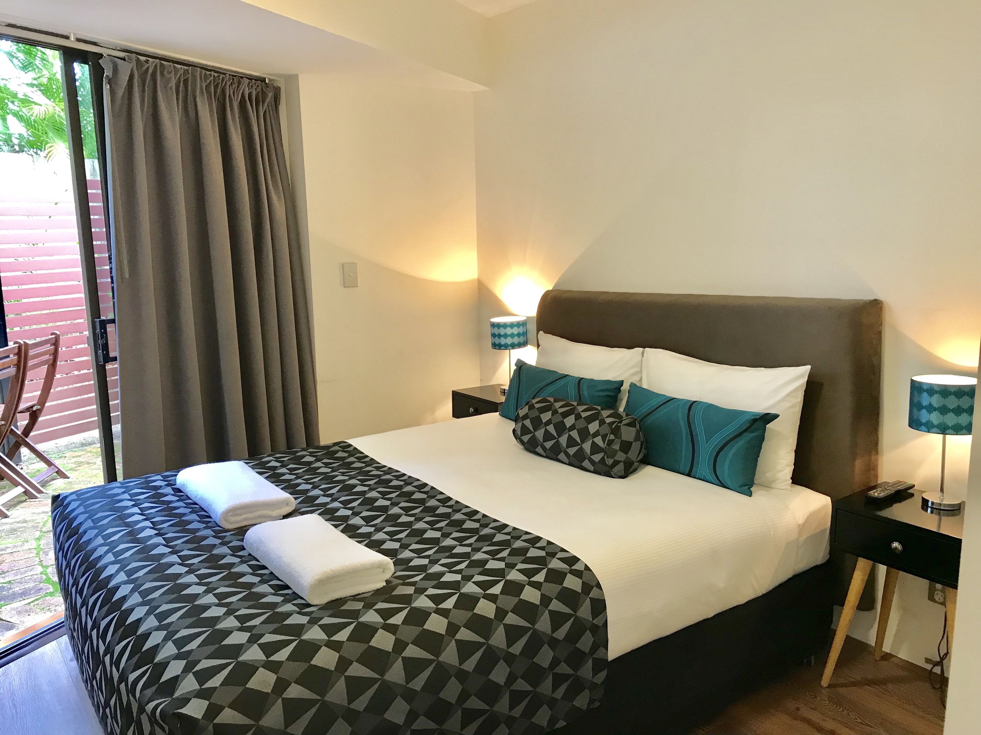 A neatly made bed with a geometric-patterned bedspread, two pillows, and two folded towels. There are two bedside lamps with patterned shades, a small bedside table, and a sliding glass door leading to an outdoor area with a wooden chair.
