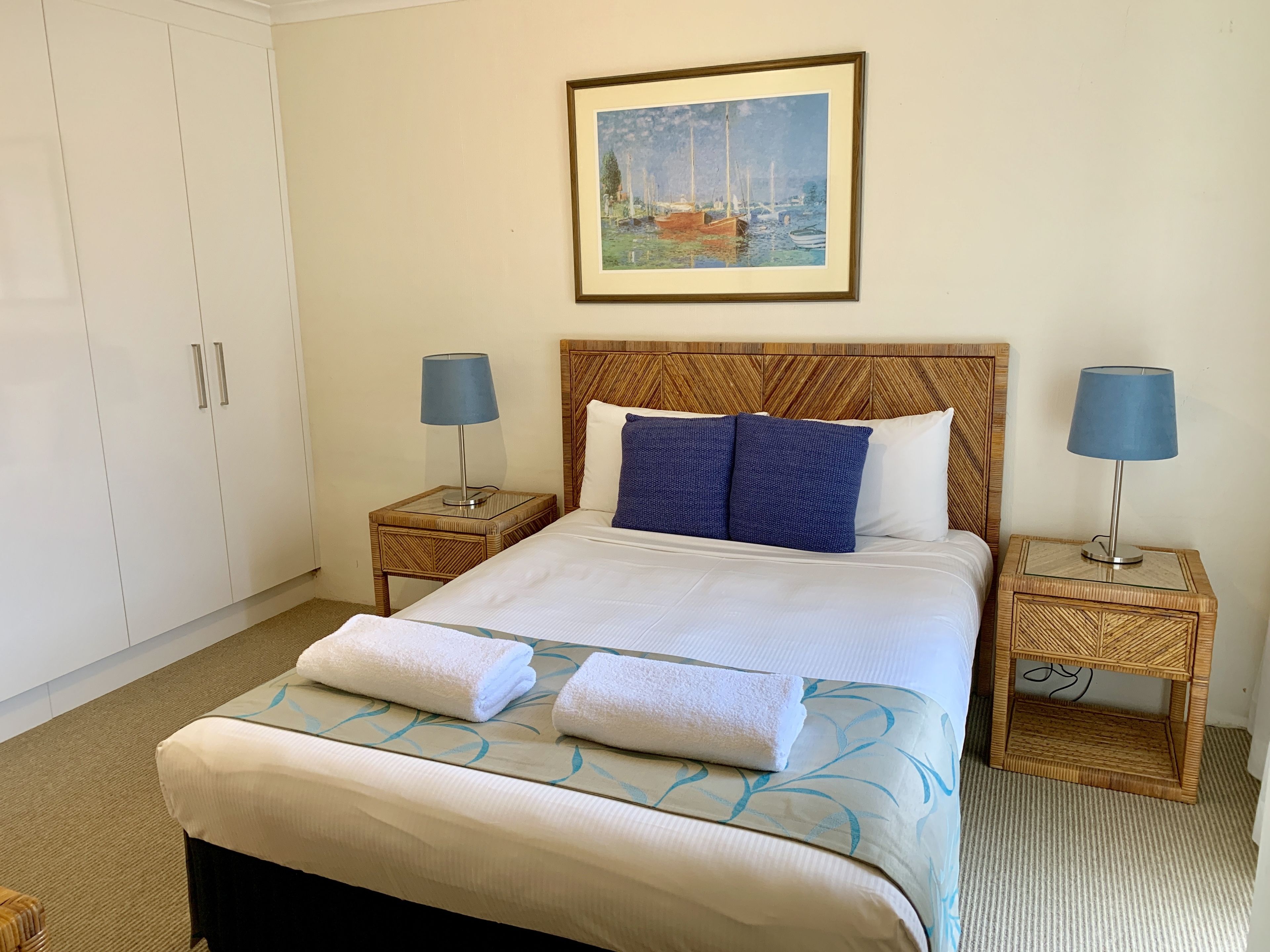 A neatly made bed with white linens and blue pillows, flanked by two nightstands with lamps, in a room with a large window and a framed painting above the bed.