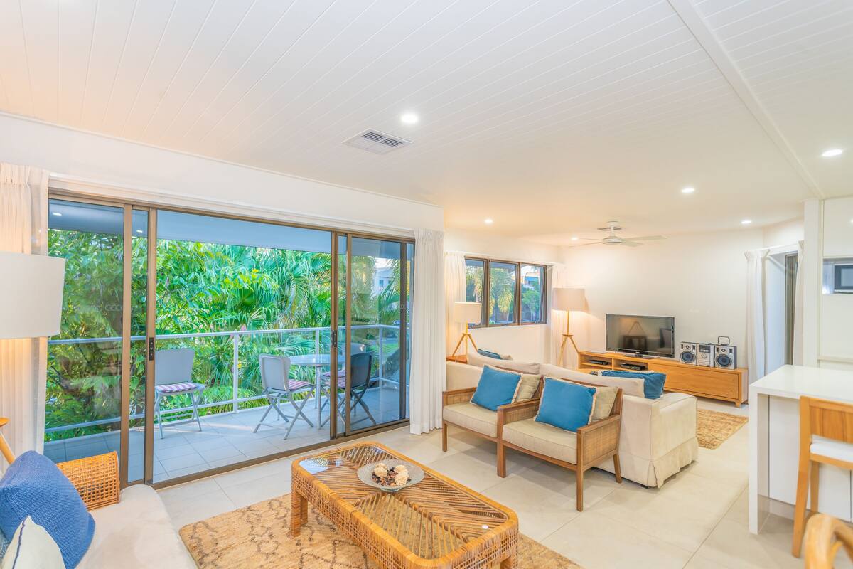 A modern living room with a sofa, coffee table, and TV. There is a balcony with glass doors and a view of greenery.