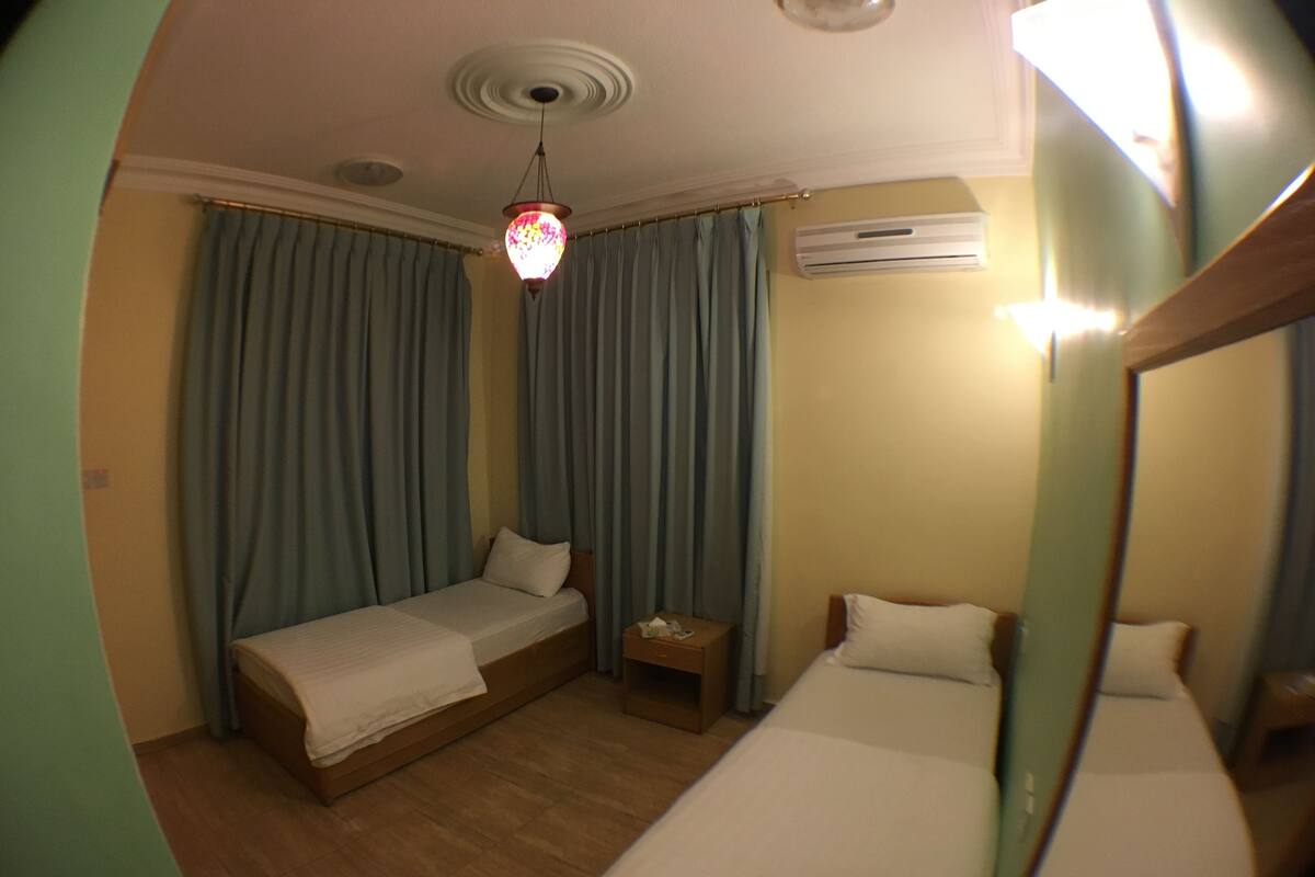 A room with two single beds, a small table, an air conditioning unit, and a ceiling light fixture.