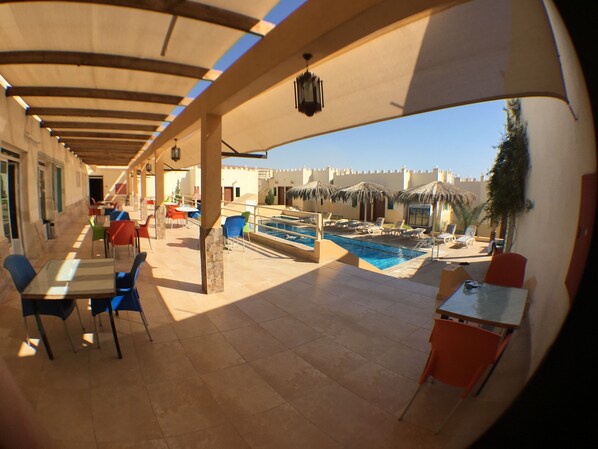 Zona de piscina al aire libre con sillones de descanso y mesas, un espacio de estar cubierto y vistas a la piscina.