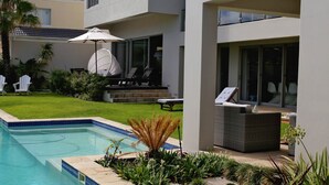 A modern outdoor pool area with a clear blue pool, surrounded by a well-maintained lawn and various plants. There are lounge chairs and a dining table visible, suggesting a space for relaxation and dining. - Da Heim Guest House (Cape Town)