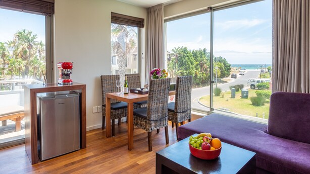 A modern hotel room with a dining area, a kitchenette, and a large window offering a view of the sea and greenery. - Da Heim Guest House (Cape Town)