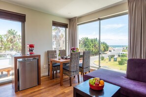 Een moderne hotelkamer met een zithoek, een kitchenette en een groot raam met uitzicht op zee en groen.