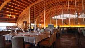 Uma ampla sala de jantar com tetos de madeira alta e uma parede de pedra. Mesas montadas com copos de vinho e pratos.
