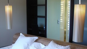 Cama bem arrumada com lençóis e almofadas brancas, telefone ao lado e um espelho refletindo um quarto com porta e cadeira.
