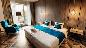 Un cuarto de hotel con una cama grande, un sillón azul, una mesita de noche y vistas al exterior a través de una ventana.