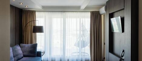 Een moderne hotelkamer met een bank, een aan de muur bevestigde televisie en een groot raam met dunne gordijnen.