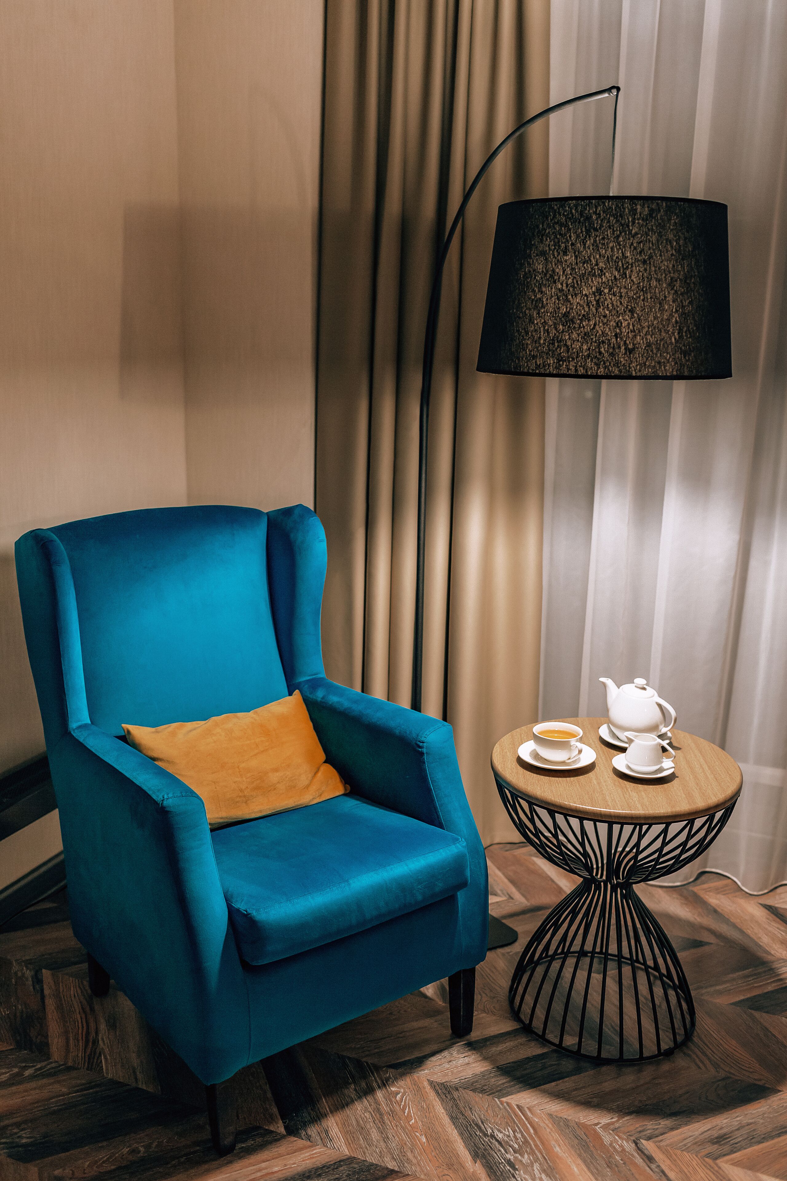 Un fauteuil bleu avec un coussin en bois, une petite table ronde avec une théière et des tasses, et un lampadaire.