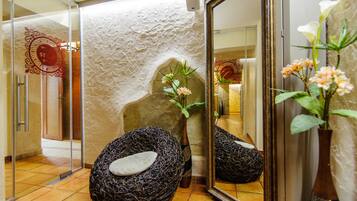 Une salle de spa avec un mur à texture, un banc en pierre, un vase rempli de fleurs et un miroir qui reflète la pièce.