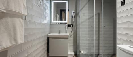 Un baño moderno con paredes de azulejos blancos, cabina de ducha con mampara de vidrio, lavamanos blanco y un espejo con luz superior.