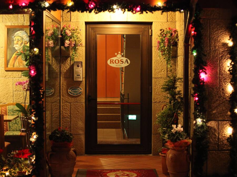 The entrance of the Rosa Hotel with a wooden door, a sign above it, and potted plants around the entrance.