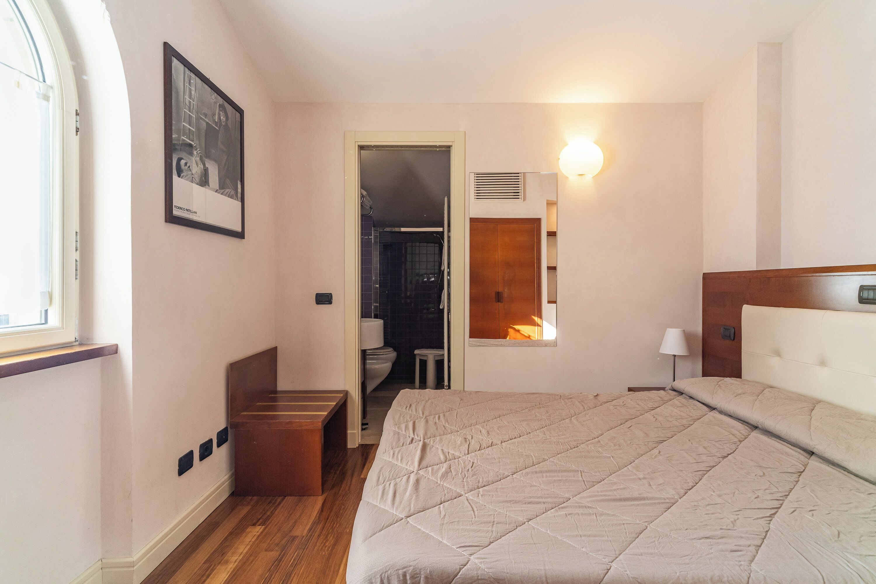 A bedroom with a bed, a wooden bench, a framed picture on the wall, and a bathroom visible through an open door.