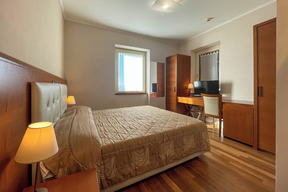 A bedroom with a bed, bedside lamp, wooden headboard, a desk with a chair, a television, and a wardrobe.
