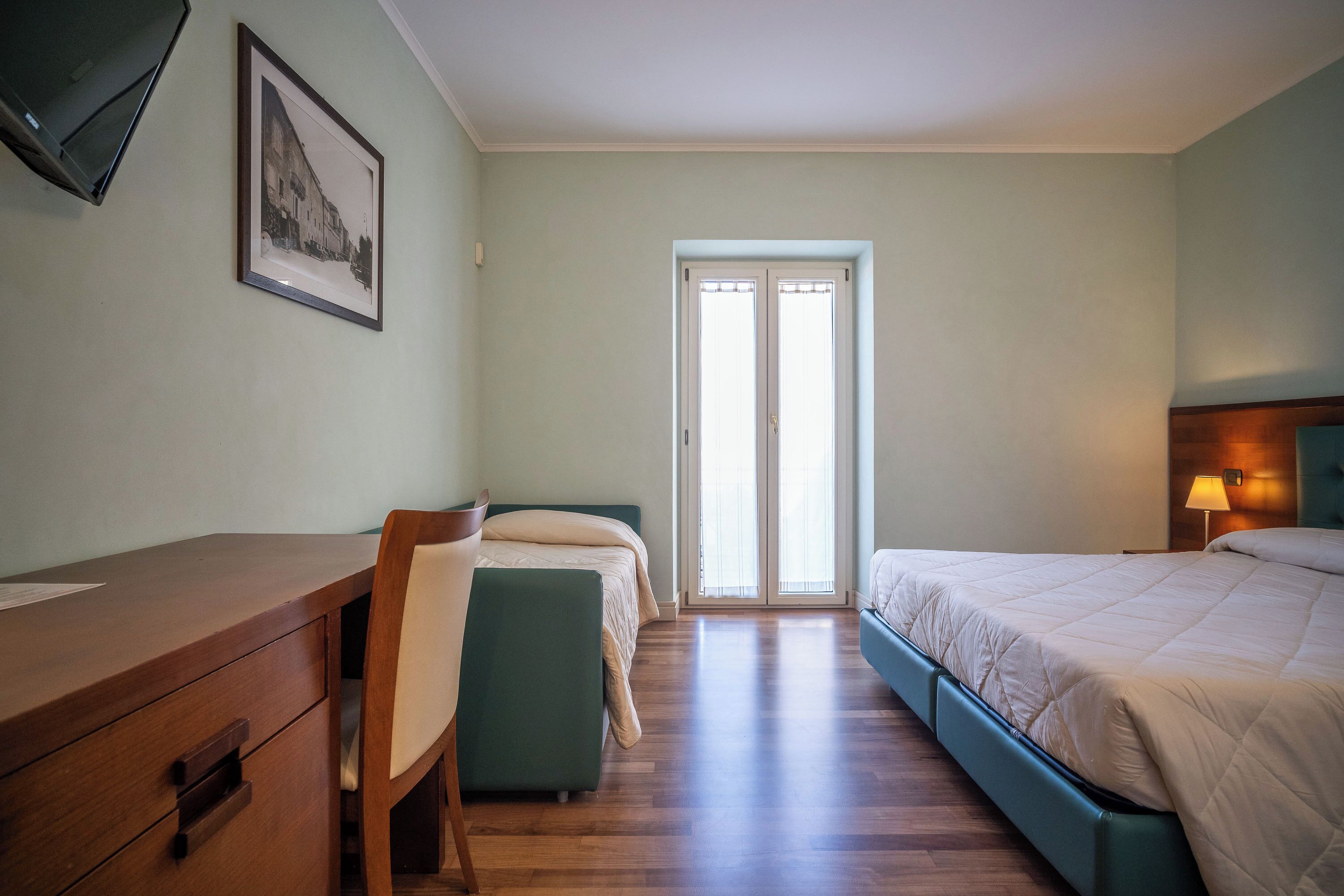 Een slaapkamer met een bureau, twee bedden, een stoel en een ingelijste foto aan de muur.
