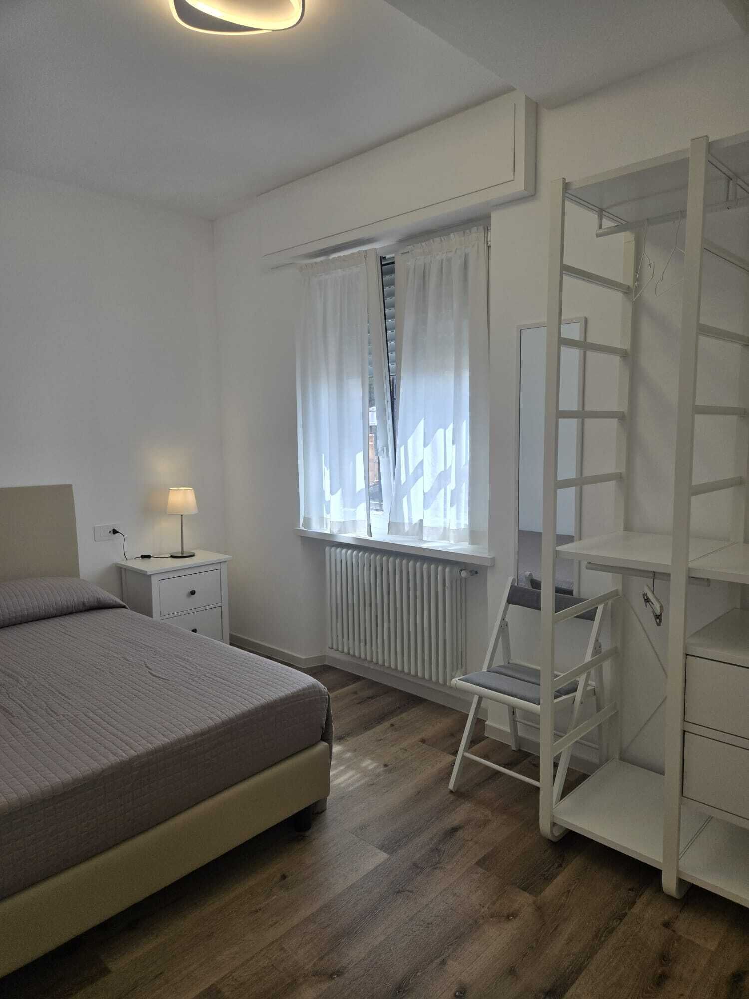 Un dormitorio con cama, mesita de noche, lámpara, ventana con cortinas, radiador y un armario blanco.