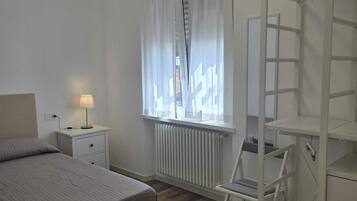 Un dormitorio con cama, mesita de noche, lámpara, ventana con cortinas, radiador y un armario blanco.