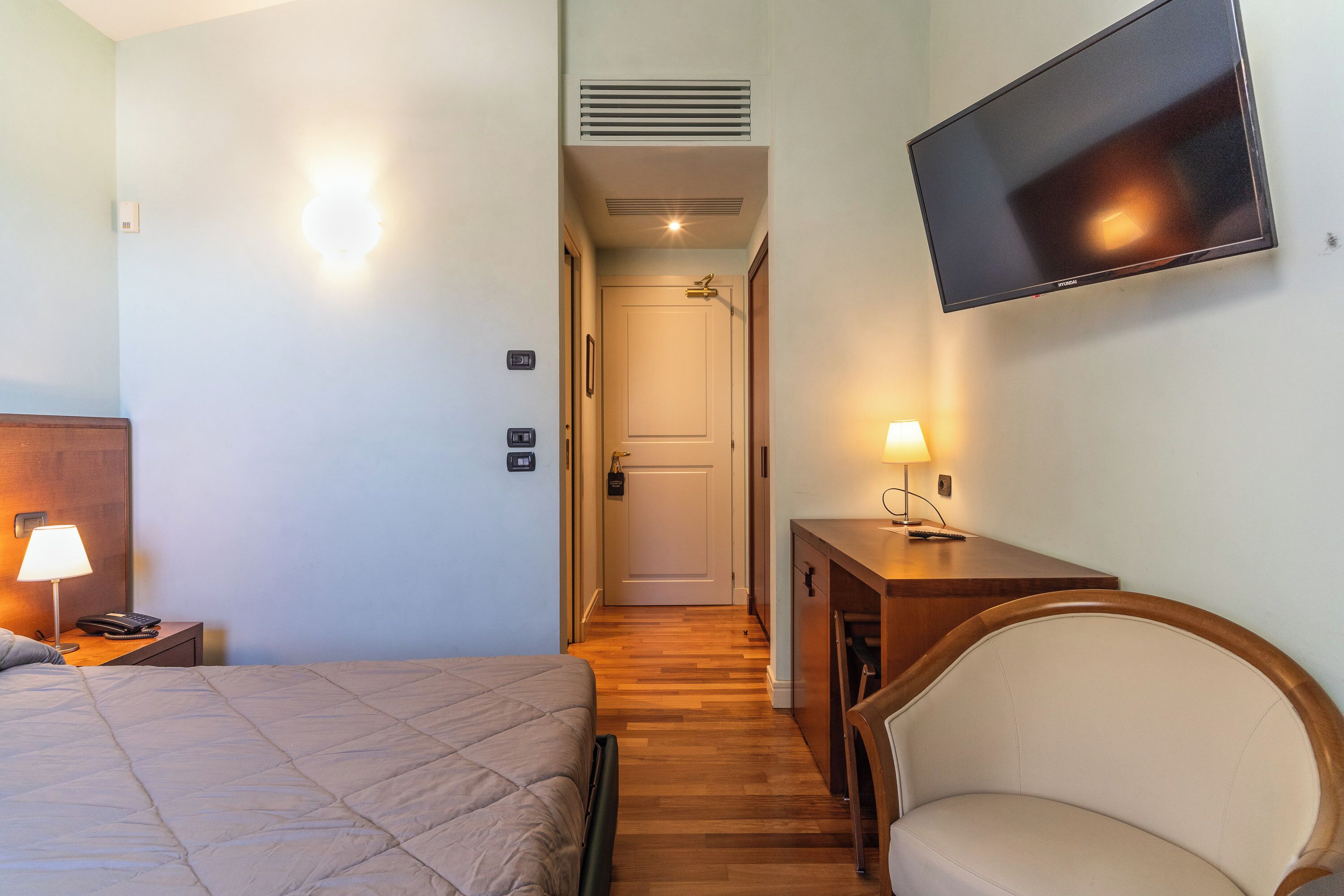 A hotel room with a bed, bedside table, lamp, TV mounted on the wall, and a chair.