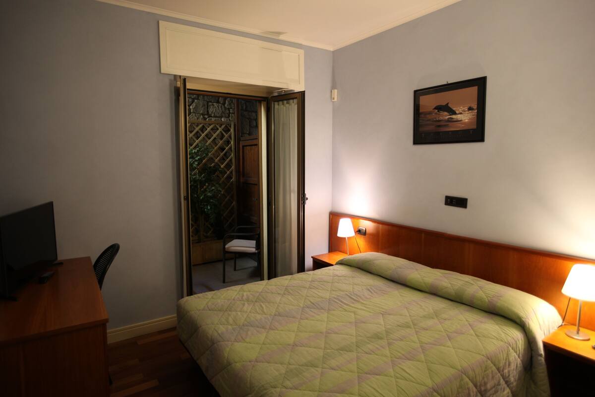 A hotel room with a bed, a desk with a computer, a chair, a nightstand with a lamp, and a door leading to a balcony.