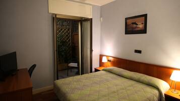 Ein Hotelzimmer mit Bett, Schreibtisch mit Computer, Stuhl, Nachttisch mit Lampe und einer Tür zu einem Balkon.