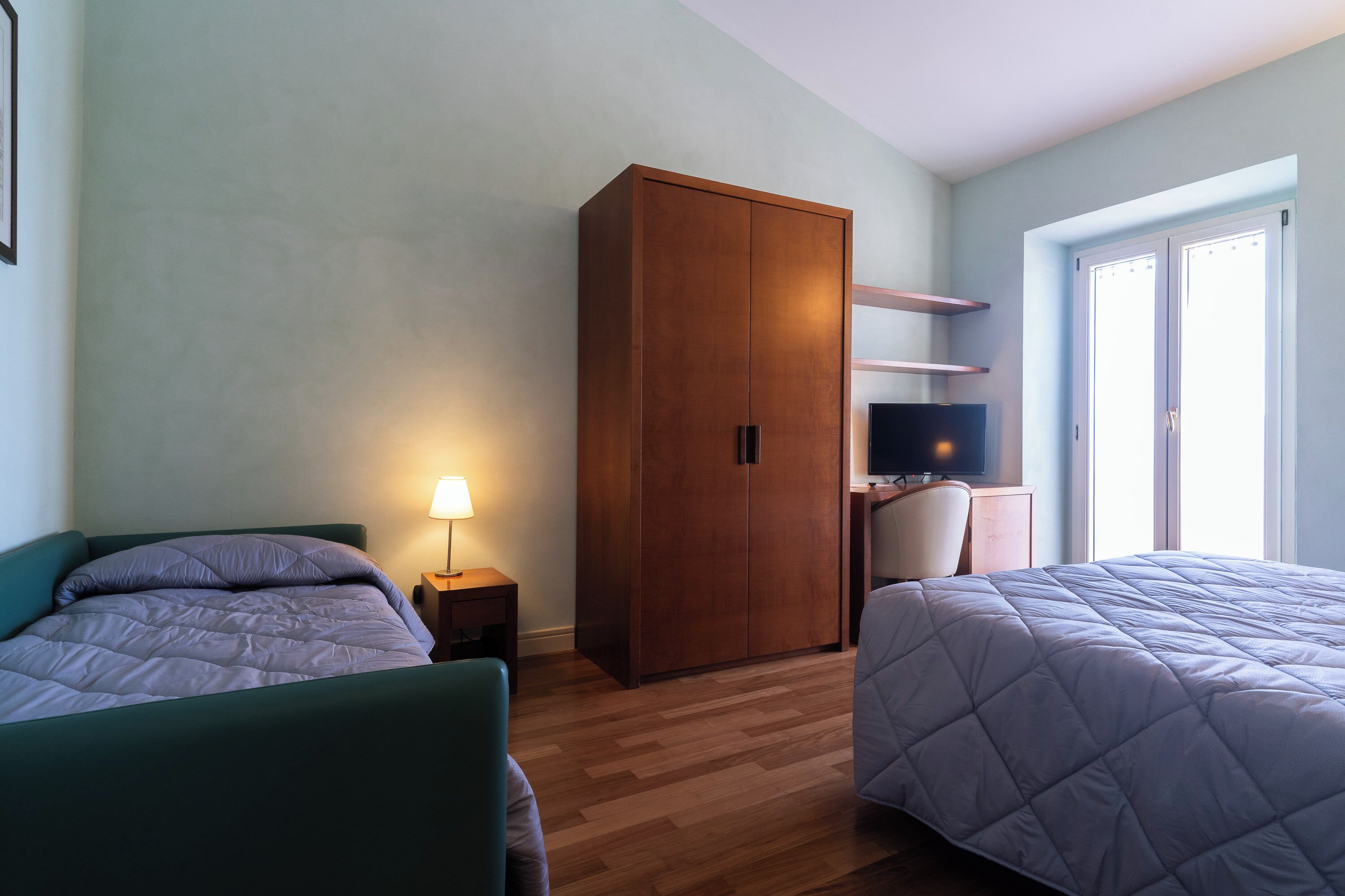 A hotel room with two beds, a wardrobe, a desk with a chair, a TV, and a window with blinds.
