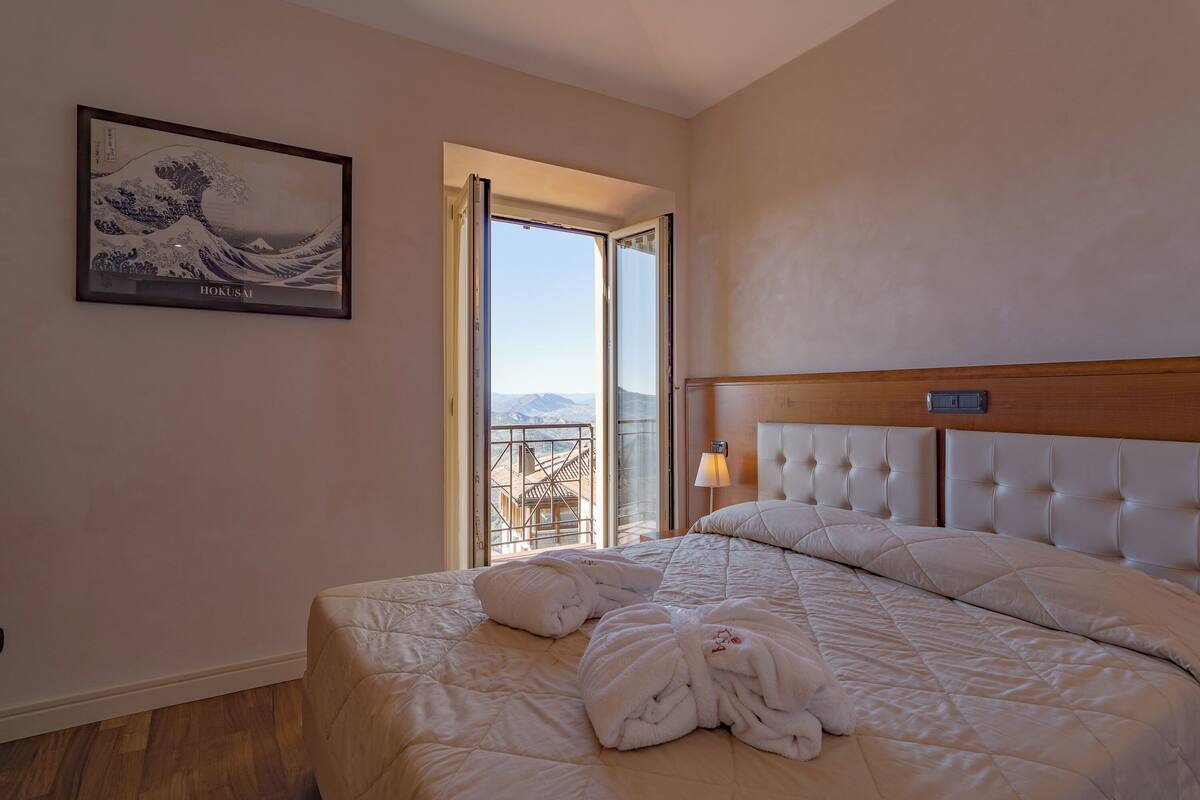 A hotel room with a large bed, a view of mountains through an open door, and a framed artwork on the wall.