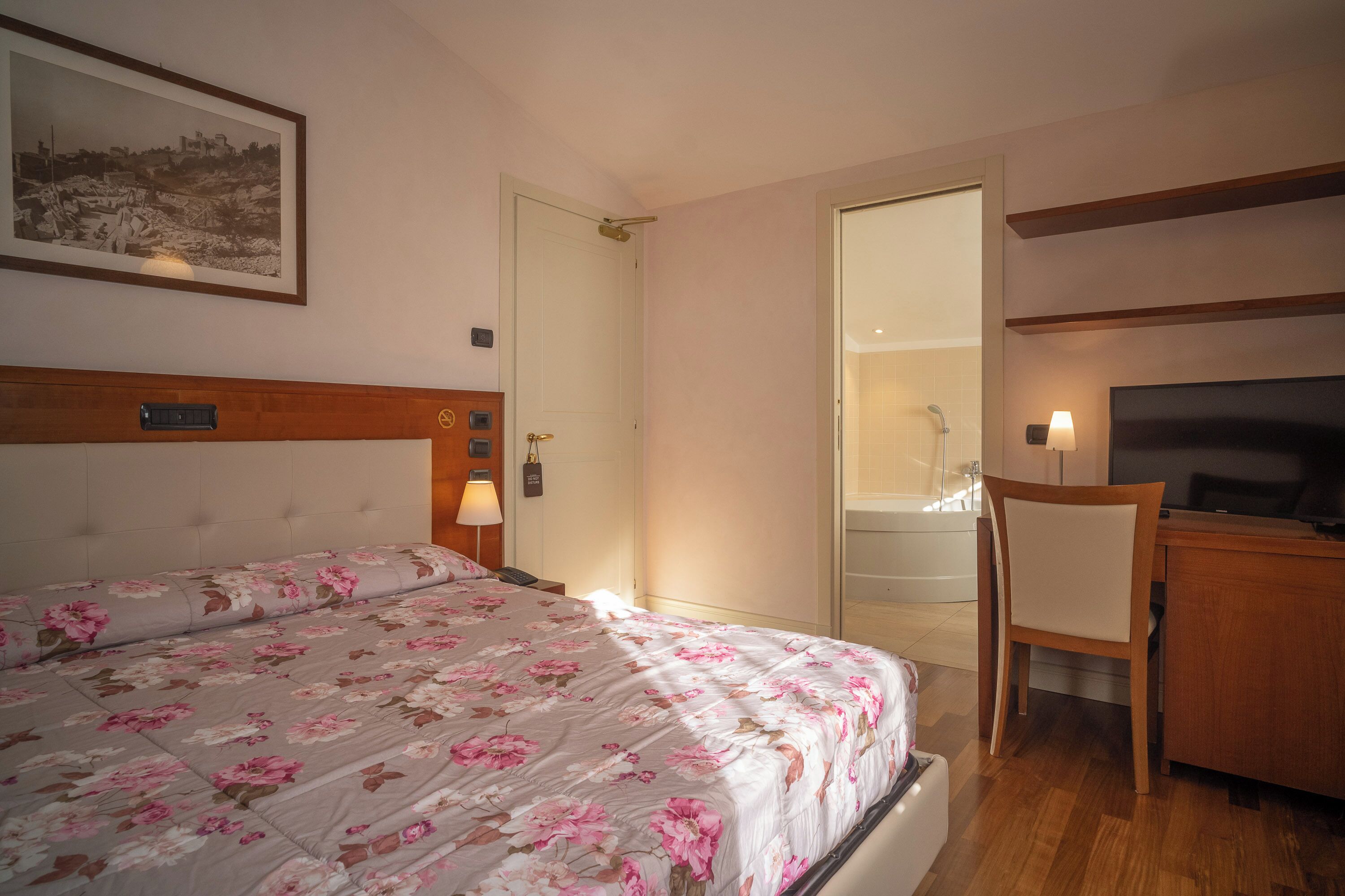 A hotel room with a bed, a desk, a chair, a television, and a bathroom visible through an open door.