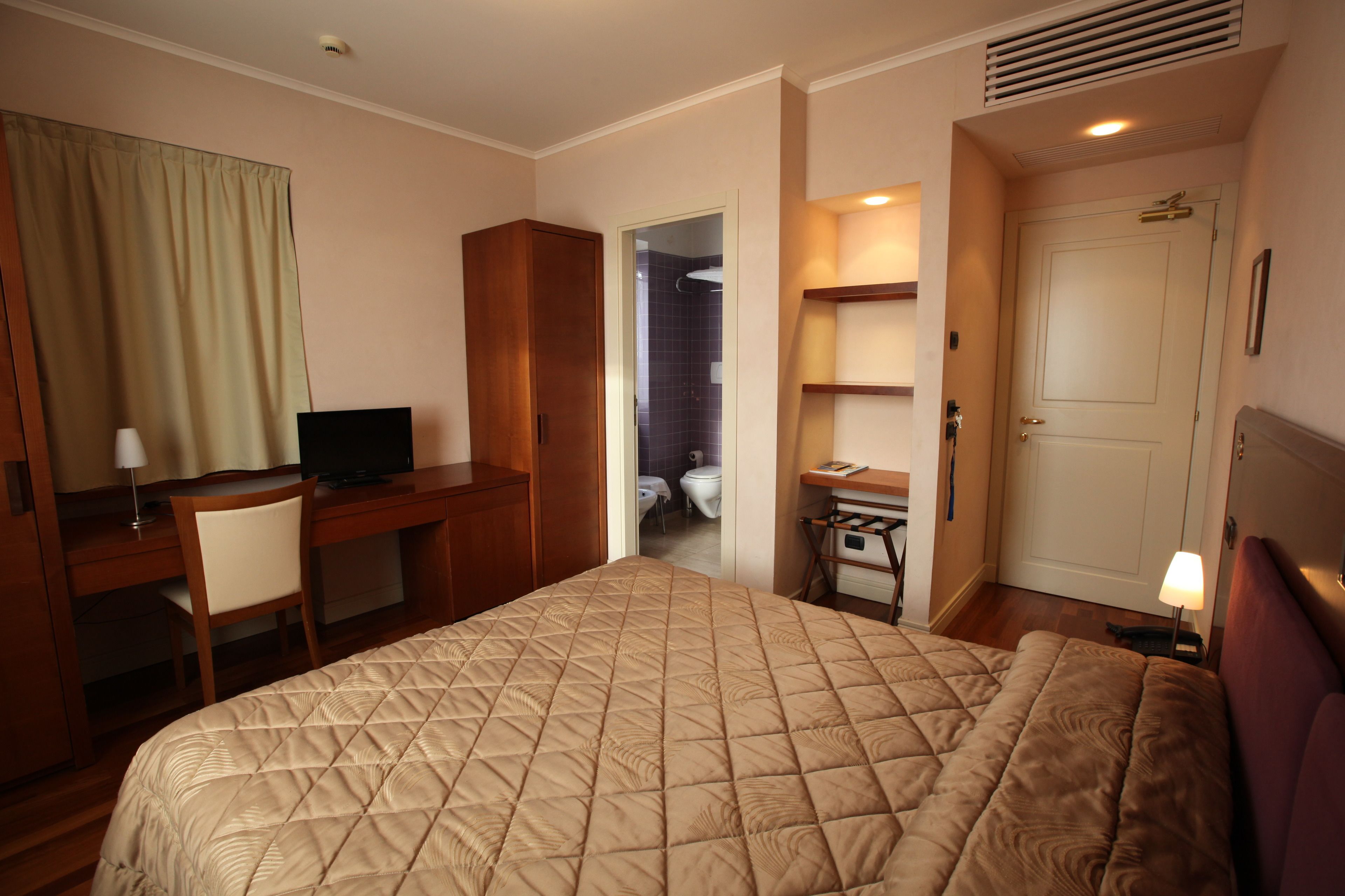 Ein Hotelzimmer mit einem großen Bett, einem Schreibtisch mit Stuhl, einem Fernseher, einem durchgegangenen Badezimmer und einem kleinen Tisch mit Lampe.