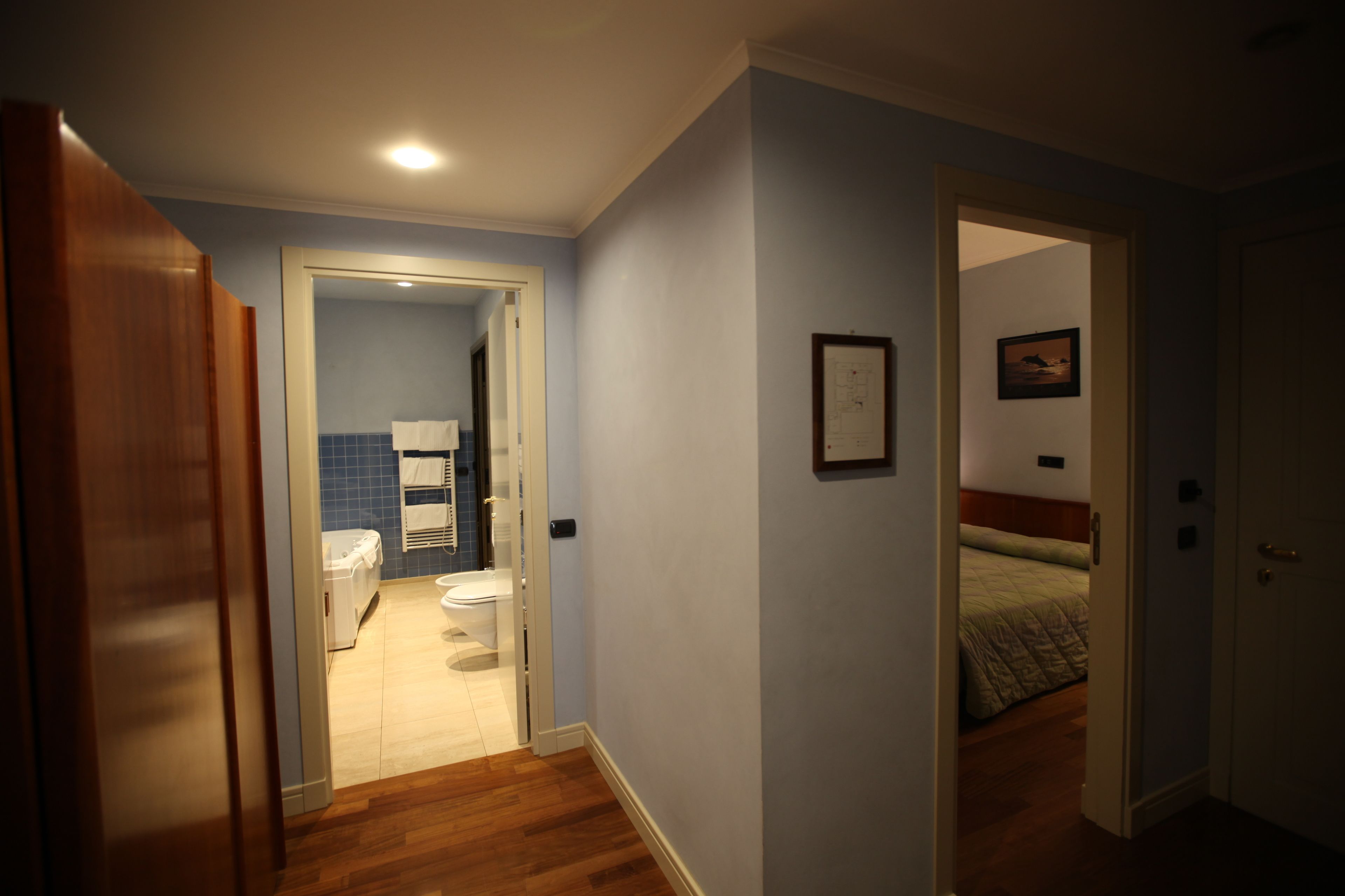 Un pasillo que da a un baño con inodoro y lavamanos, un dormitorio con cama, y una puerta que conduce a otra habitación.