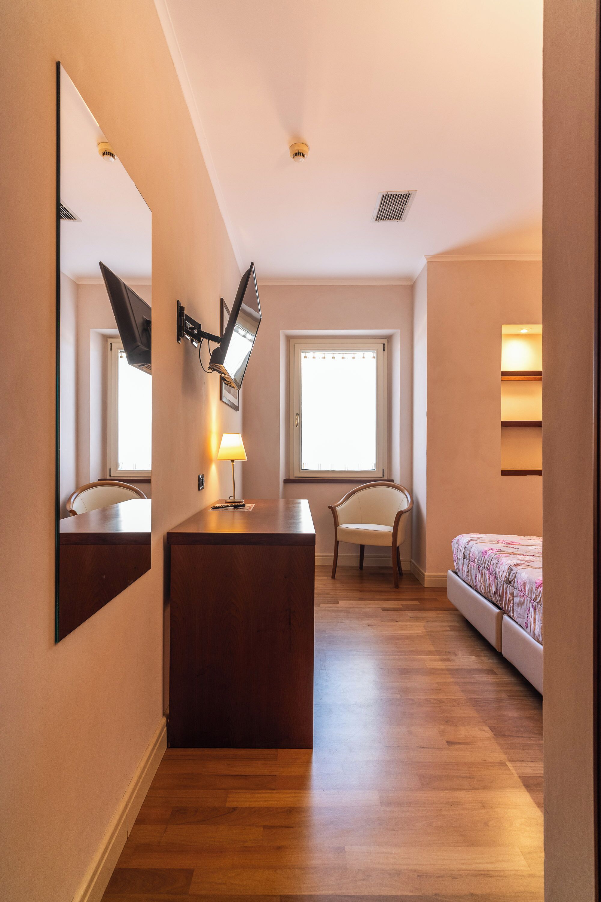 A hotel room with a wooden desk, a chair, a bed, a large mirror, and a window with blinds.