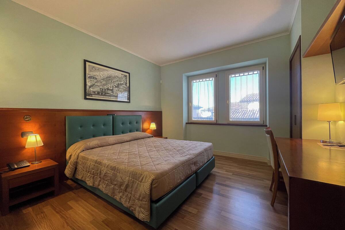 A bedroom with a bed, bedside lamps, a framed picture on the wall, a window with curtains, and a wooden desk with a lamp.