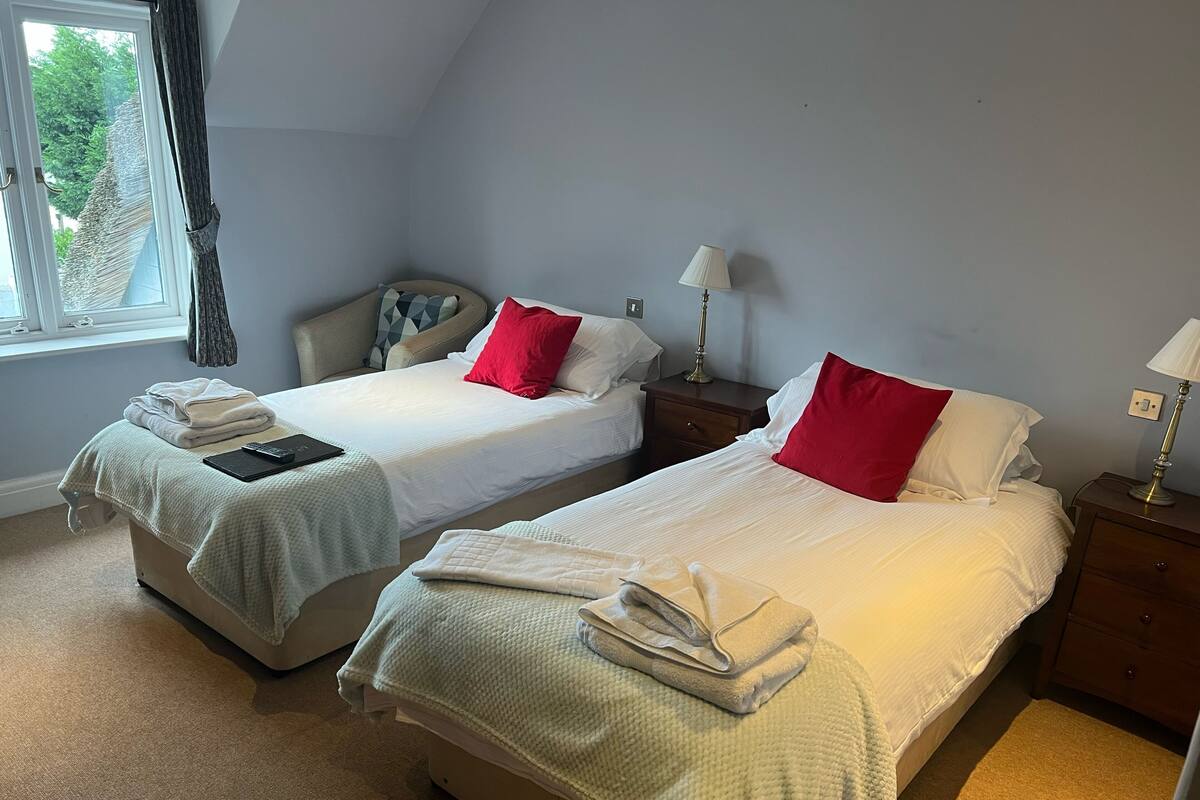 A room with two beds, each with a white bedspread and red pillows, a bedside table with a lamp, and a window with curtains.
