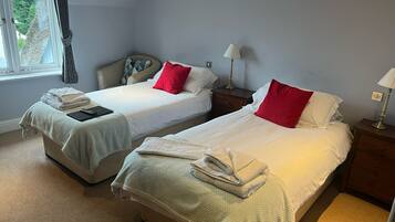 A room with two beds, each with a white bedspread and red pillows, a bedside table with a lamp, and a window with curtains.