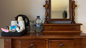A wooden desk with a mirror, two drawers, a small stool, and various personal items including a water bottle, coffee maker, and snacks.
