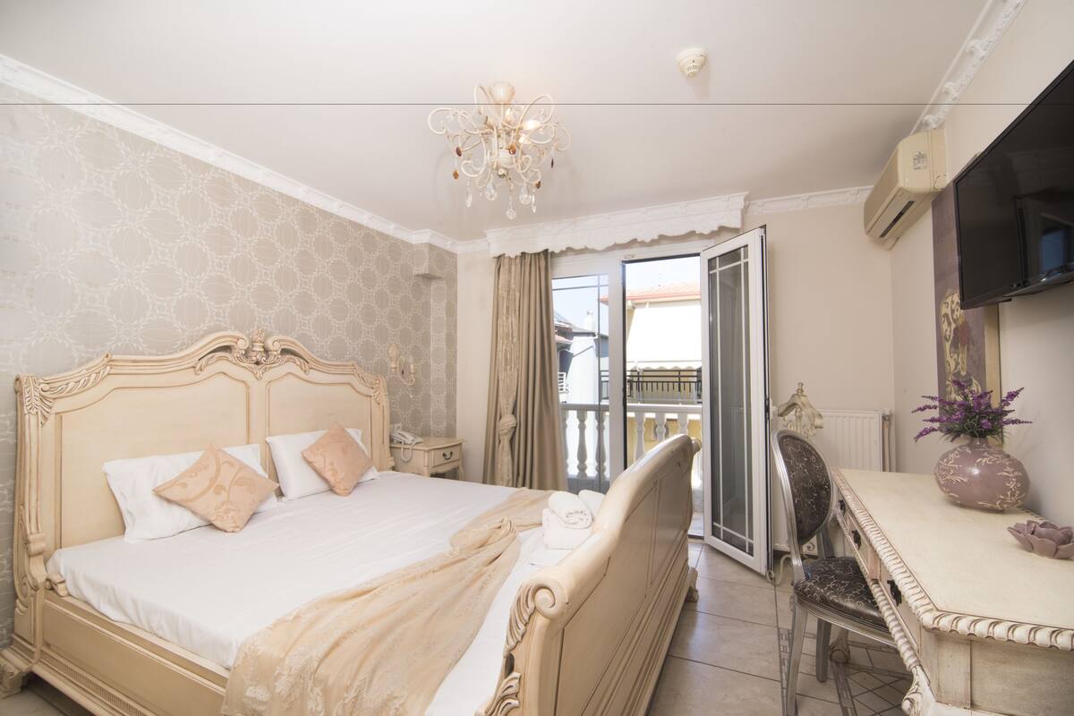Quarto com uma cama grande, uma escrivaninha e uma cadeira. Há um candelabro, uma televisão e uma janela com cortinas.