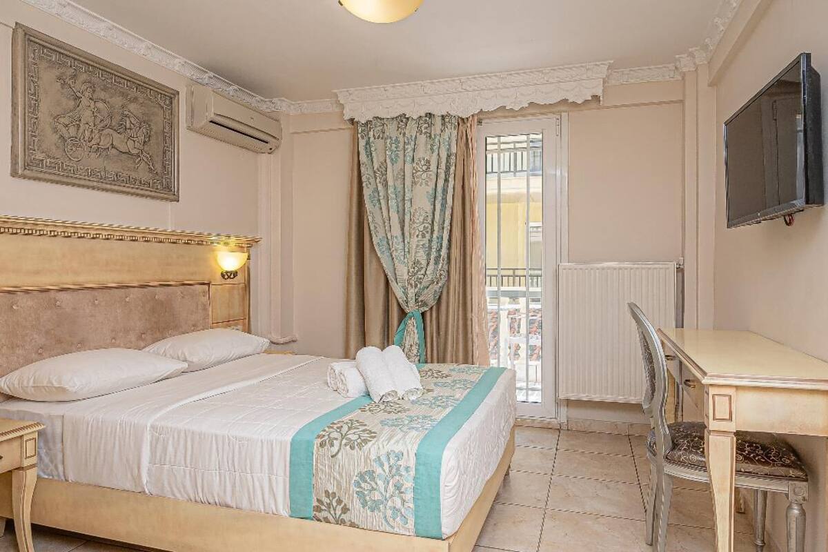 Quarto com cama, escrivaninha, cadeira, televisão e janela com cortinas.