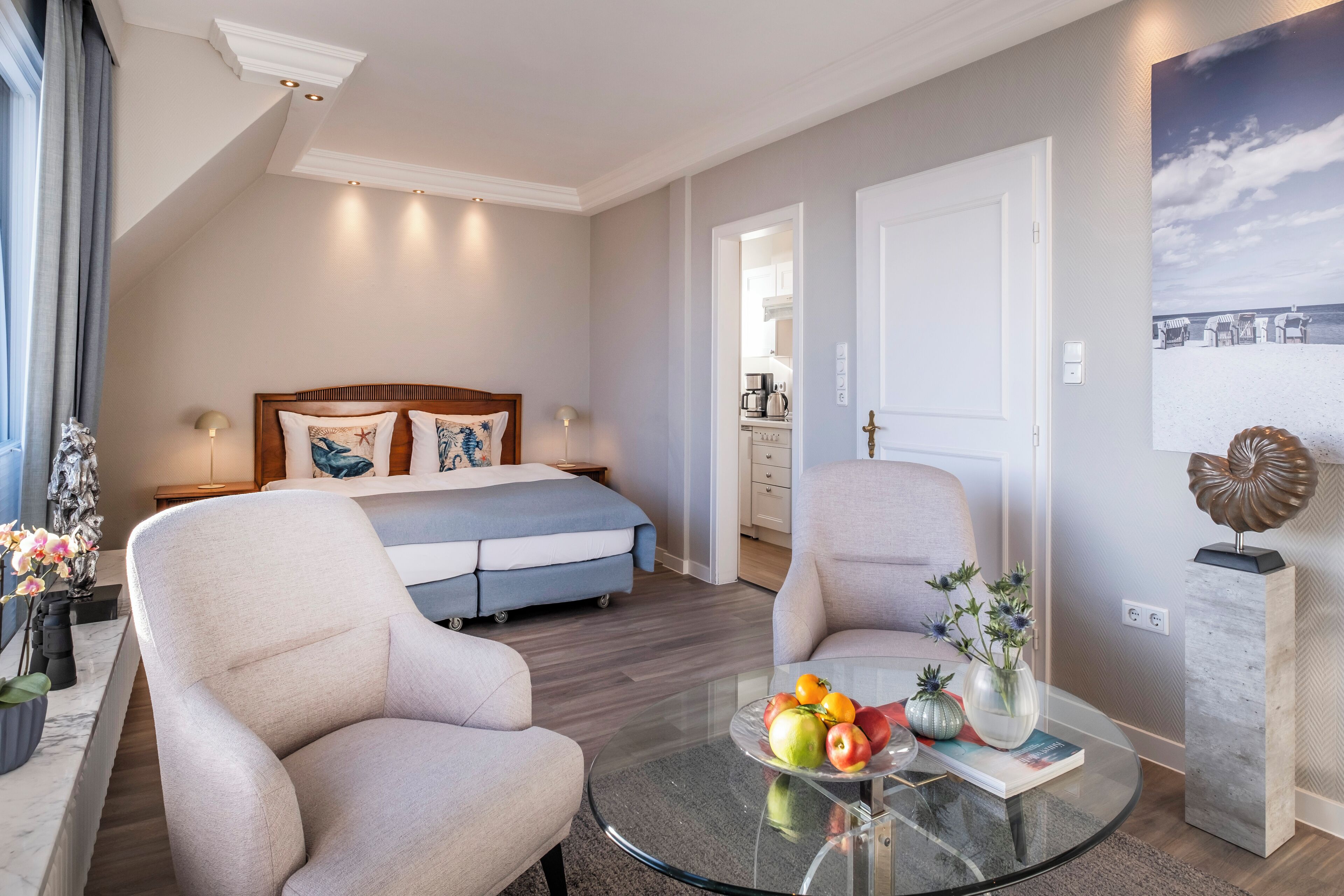 Ein modernes Hotelzimmer mit einem Bett, zwei Sesseln, einem Glastisch mit Früchten und einem Bild einer Strandansicht.