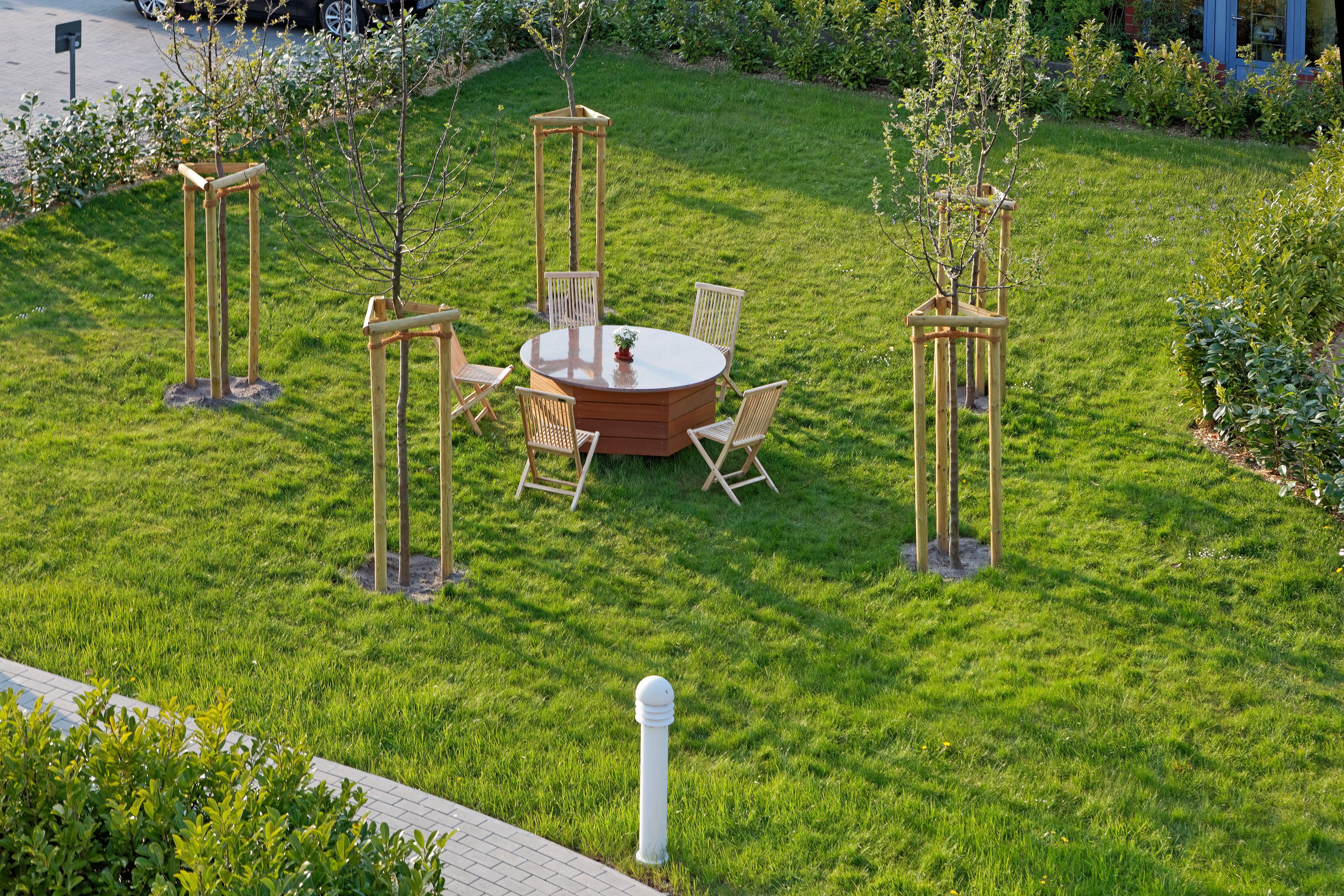 A well-maintained outdoor seating area with a round table and chairs, surrounded by small trees and a grassy lawn.