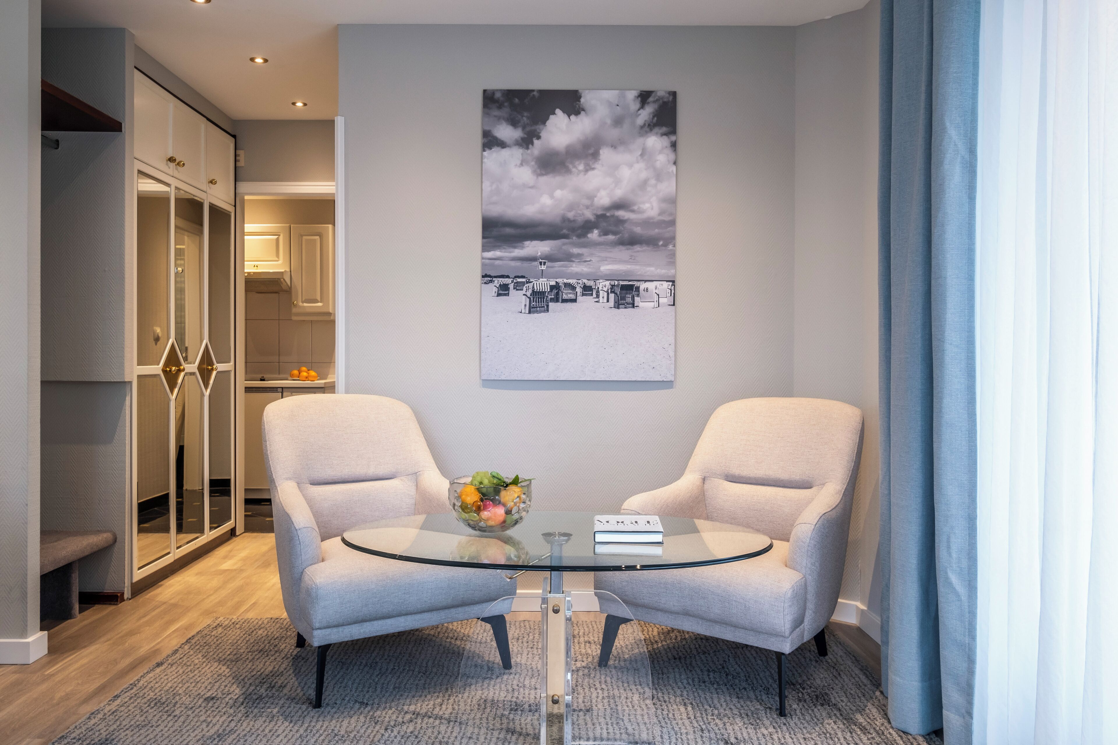 Ein modernes Zimmer mit zwei Sesseln, einem Glastisch mit Obstschale und einem Schwarz-Weiß-Foto an der Wand.
