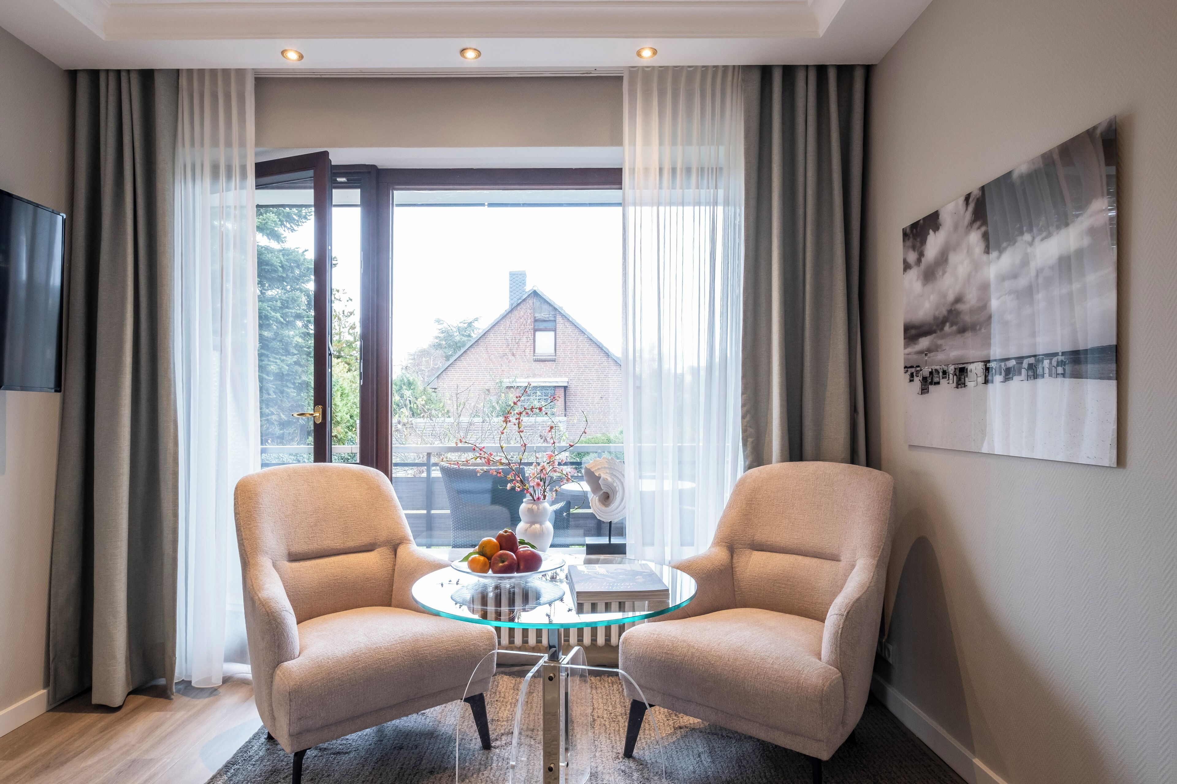 Una habitación moderna con dos sillones color beige, una mesa de centro de cristal con frutero, y vista a un edificio de ladrillo a través de amplias ventanas.