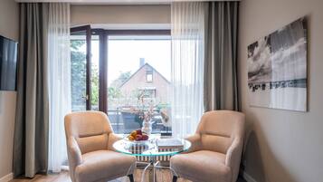 A modern room with two beige armchairs, a glass coffee table with a fruit bowl, and a view of a brick building through large windows.